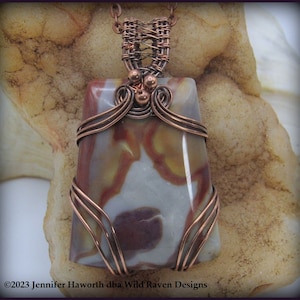 May include: A handcrafted pendant featuring a rectangular stone with a mix of gray, brown, and cream colors. The stone is wrapped in copper wire, with intricate weaving at the top and decorative swirls. The pendant hangs from a copper chain.