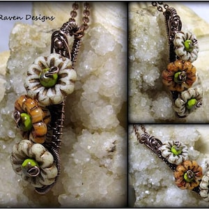 May include: A copper wire wrapped necklace with three ceramic flowers. The flowers are white with green and orange accents. The necklace is on a white background.