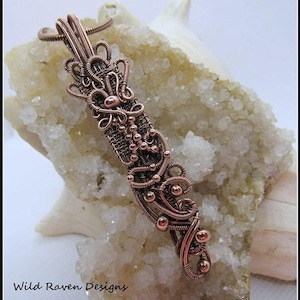 Puede incluir: Un collar con colgante de alambre de cobre con intrincados diseños tejidos y pequeñas cuentas de cobre. El colgante se muestra sobre una roca blanca con textura cristalina.