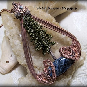 Pode incluir: Um pingente envolto em fio de cobre com uma pedra de ágata azul e branca, uma árvore envolvida em fio verde e o texto "Wild Raven Designs".