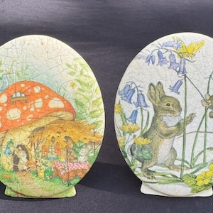 Può includere: Due sottobicchieri in ceramica ovali con un tema boschivo fantasioso. Uno presenta un fungo rosso e bianco con una piccola casa in cima e un gruppo di animali riuniti intorno a un tavolo da picnic. L'altro presenta un coniglio marrone e un topo che giocano su una vite con campanule blu e fiori gialli.