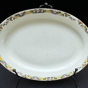 May include: Oval white serving platter with a decorative border. The border features a yellow band with pink and yellow floral accents. The platter is supported by a black metal stand.