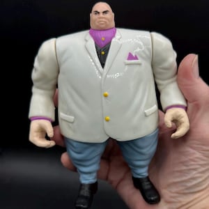May include: A Kingpin action figure, featuring a white suit with yellow buttons, a purple ascot, and blue pants. The figure has a stern expression and black shoes. The figure is held in a hand.
