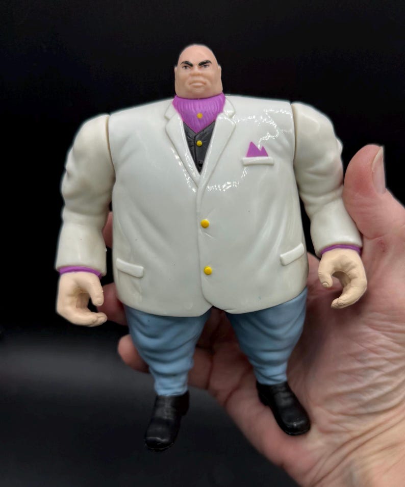 May include: A Kingpin action figure, featuring a white suit with yellow buttons, a purple ascot, and blue pants. The figure has a stern expression and black shoes. The figure is held in a hand.