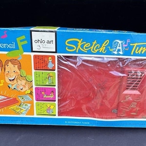 May include: Vintage "Sketch A Tune" musical pencil toy in its original box. The box is blue with colourful illustrations and text, including "Electronic Magic Musical Pencil" and "24 Different Tunes". The toy itself is red.