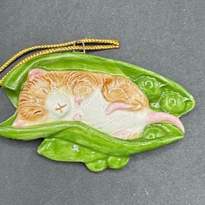 Puede incluir: Adorno de cerámica con un hámster durmiendo en una vaina de guisantes verde. El hámster tiene pelaje marrón y blanco con detalles rosas. Una cuerda dorada está unida para colgarlo. La vaina de guisantes es de color verde brillante.