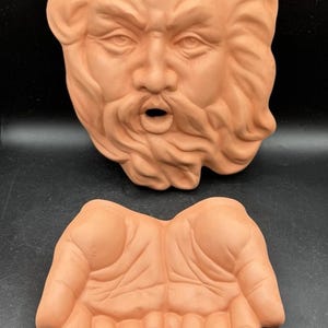 Könnte beinhalten: Terrakotta-Skulptur eines bärtigen Gesichts mit offenem Mund und einem Paar offener Hände. Das Gesicht hat detaillierte Merkmale und die Hände sind gewölbt, was auf einen Behälter hindeutet. Die Objekte sind in einem warmen, erdigen Ton gehalten.
