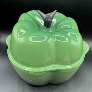 Puede incluir: Una cocotte Le Creuset verde de hierro fundido, con forma de pimiento. La tapa tiene un asa negra en forma de tallo. La cocotte tiene un acabado brillante y dos asas laterales. La forma general es un diseño redondeado, similar a una verdura.
