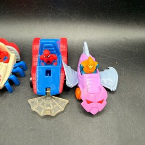 May include: Three toy vehicles featuring Spider-Man and the Green Goblin. The Spider-Man vehicle is a red and blue tank-like car. The Green Goblin vehicle is a pink and purple glider. A clear plastic spiderweb is in front.