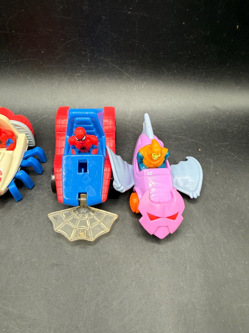 May include: Three toy vehicles featuring Spider-Man and the Green Goblin. The Spider-Man vehicle is a red and blue tank-like car. The Green Goblin vehicle is a pink and purple glider. A clear plastic spiderweb is in front.