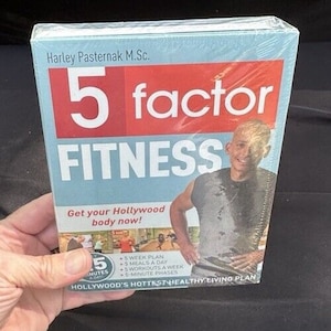 Puede incluir: Un libro titulado "5 factor FITNESS" de Harley Pasternak M.Sc. La portada presenta a un hombre con un tanque gris y el texto "¡Consigue tu cuerpo de Hollywood ahora!"  La portada del libro también incluye un plan de entrenamiento de 5 minutos, 5 comidas al día, 5 entrenamientos a la semana y fases de 5 minutos. El libro se describe como "El plan de vida saludable más popular de Hollywood."