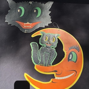 May include: Two vintage Halloween decorations. One is a black cat face with green eyes and a red mouth. The other is a black cat sitting on an orange crescent moon with a smiling face. Both have string for hanging.
