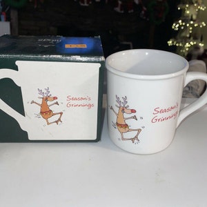 Könnte beinhalten: Weiße Keramiktasse mit einer roten Rentier-Illustration und dem Text "Season's Grinnings".