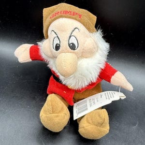 May include: A plush Grumpy character doll from Snow White wearing a brown hat with the word GRUMPY embroidered in red, a red tunic, and brown pants. The doll features a white faux fur beard and has a white paper tag attached.