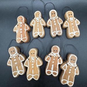 May include: Eight wooden gingerbread man ornaments with white icing details and black wire hangers. The ornaments are brown with white icing accents, including buttons, bows, and facial features. These festive decorations are perfect for Christmas.