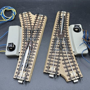 May include: Two brown and tan model train track pieces with silver metal switches. The track pieces are connected to a gray electrical switch box with wires.