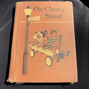 Könnte beinhalten: Vintage-Buch mit dem Titel "On Cherry Street" mit einer Illustration von Kindern in einem Wagen. Das Cover ist in verblichenem Orange gehalten und zeigt eine Straßenlaterne und die Worte "Cherry St."