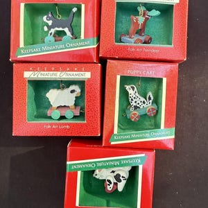 May include: Five miniature keepsake ornaments in red boxes. Each box features a different animal on a cart: a black and white cat, a reindeer, a lamb, a dalmatian, and a bunny. The boxes have green interiors and text that reads "Kitty Cart", "Puppy Cart", and "Folk Art".
