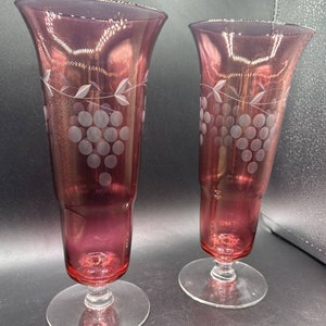 May include: Two clear glass goblets with a grapevine design etched into the glass. The goblets are a deep burgundy red color and have a clear glass base.