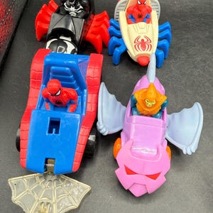 May include: Four toy vehicles featuring Marvel characters are shown. There's a red and black Venom car, a Spider-Man boat, a red and blue Spider-Man tank, and a pink Goblin vehicle. A clear plastic spiderweb is also included.