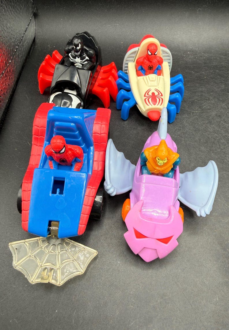 May include: Four toy vehicles featuring Marvel characters are shown. There's a red and black Venom car, a Spider-Man boat, a red and blue Spider-Man tank, and a pink Goblin vehicle. A clear plastic spiderweb is also included.