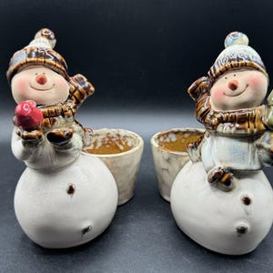 Op de afbeelding: Twee keramische sneeuwman-plantenbakken met bruine en witte verf, elk met een kleine pot. Eén sneeuwman houdt een rode ornament vast en de andere een kleine groene boom.