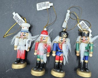 May include: Four wooden nutcracker ornaments with white hair and various outfits. One wears a chef's hat, another a Santa hat, and the others wear military-style uniforms. Each has a gold string for hanging and sits on a gold base.