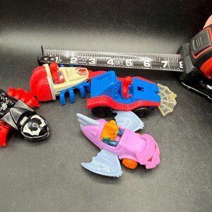 May include: A selection of colourful toy vehicles, including a red and white boat-like car, a blue and red car, a black car, and a pink airplane-shaped car. A measuring tape shows the vehicles are approximately 7.6 cm long.