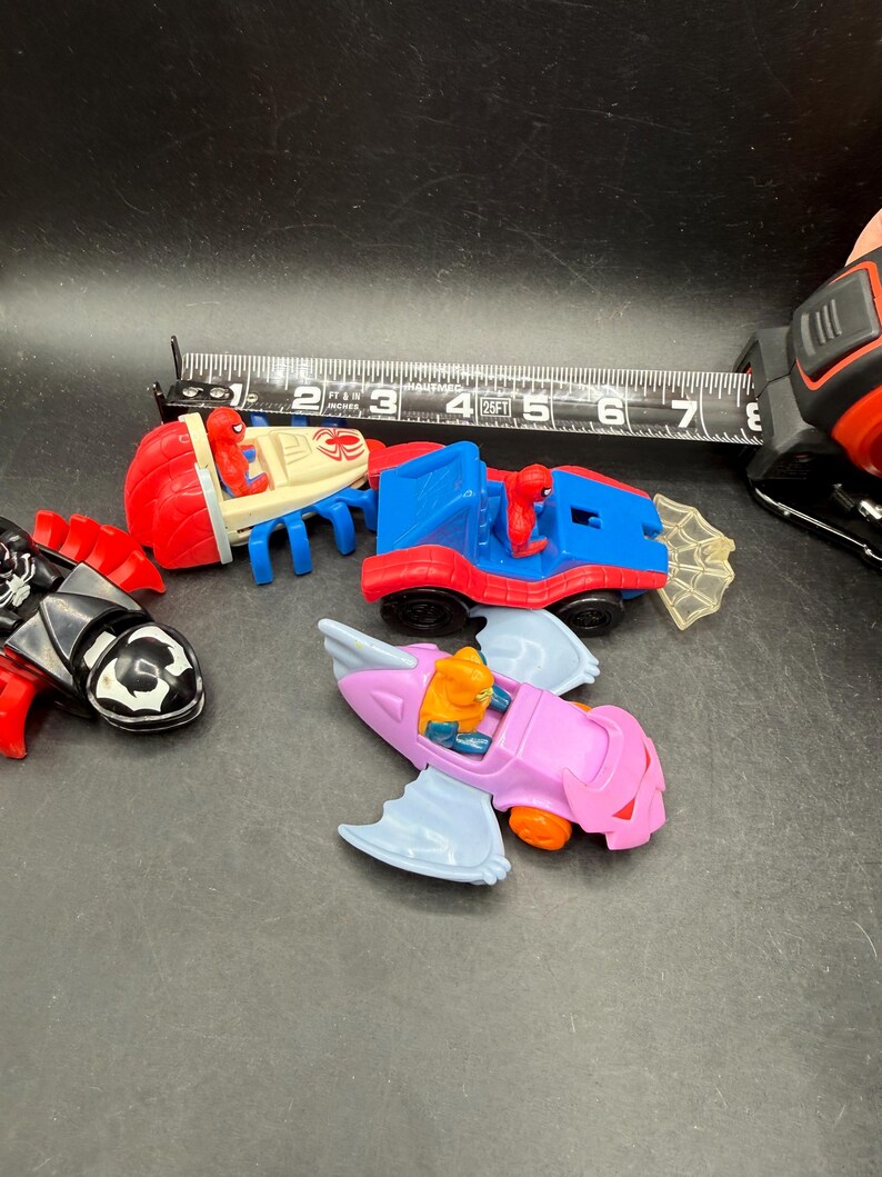 May include: A selection of colourful toy vehicles, including a red and white boat-like car, a blue and red car, a black car, and a pink airplane-shaped car. A measuring tape shows the vehicles are approximately 7.6 cm long.
