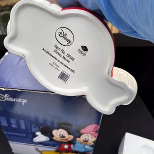 May include: A white ceramic Mickey Mouse-shaped item with red accents. The bottom of the item features the Disney logo, item details, and the text "Not dishwasher or microwave safe."