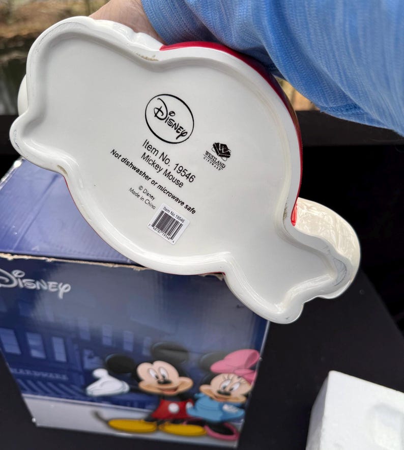 May include: A white ceramic Mickey Mouse-shaped item with red accents. The bottom of the item features the Disney logo, item details, and the text "Not dishwasher or microwave safe."