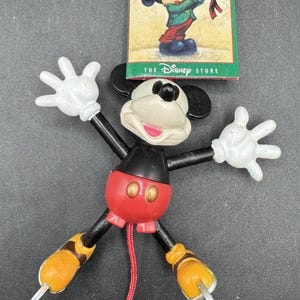 May include: A Mickey Mouse figurine with articulated limbs, featuring a red torso, black arms and legs, and white gloves. The figurine is accompanied by a tag from The Disney Store, with the text "Mickey's Season of Song".