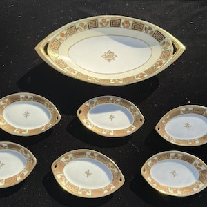 Könnte beinhalten: Ein Set aus sieben ovalen Servierplatten mit goldenen Verzierungen und einem Blumendesign. Die Platten sind weiß mit einem goldenen und braunen Muster. Die größte Platte hat zwei Griffe.