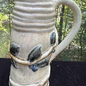 May include: A ceramic mug with a light brown glaze and a vine design with green leaves and blue berries. The mug has a handle and a rounded base.