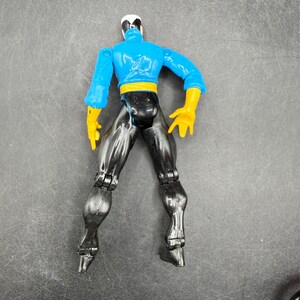 May include: A plastic action figure of a superhero, viewed from the back. The figure has a blue top with yellow gloves and a yellow belt. The pants are black, and the head is white and black.