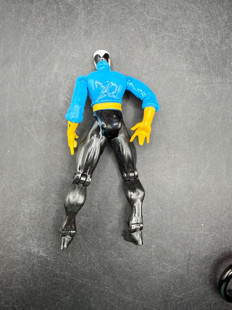 May include: A plastic action figure of a superhero, viewed from the back. The figure has a blue top with yellow gloves and a yellow belt. The pants are black, and the head is white and black.