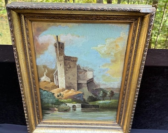 Puede incluir: Una pintura antigua enmarcada representa un castillo sobre un afloramiento rocoso, con un río en primer plano y un cielo nublado arriba. La pintura está en un marco dorado ornamentado.