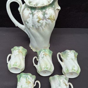 Może przedstawiać: Zestaw do herbaty z porcelany składający się z dużego dzbanka z pokrywką i pięciu pasujących filiżanek. Każdy element ma skręcony, żebrowany korpus z bladozielonym i białym opalizującym wykończeniem, ozdobiony białymi wzorami kwiatowymi i złotymi wykończeniami.