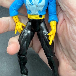 May include: A vintage action figure of a male character with a stern expression. The figure wears a blue top with a white emblem, black pants, yellow gloves, and a yellow belt. The figure is held in a hand.
