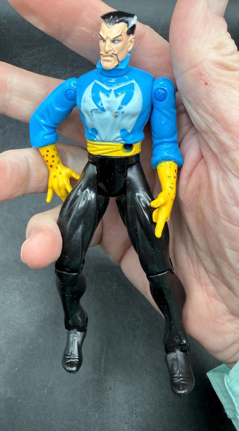 May include: A vintage action figure of a male character with a stern expression. The figure wears a blue top with a white emblem, black pants, yellow gloves, and a yellow belt. The figure is held in a hand.