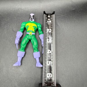 May include: A green action figure with a yellow chest and purple accents, next to a measuring tape. The figure is approximately 13 cm tall. The tape measure shows inches and feet.
