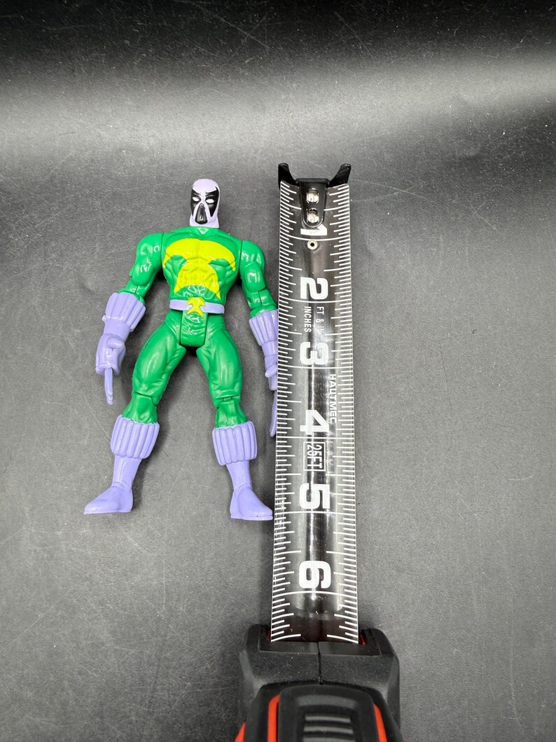 May include: A green action figure with a yellow chest and purple accents, next to a measuring tape. The figure is approximately 13 cm tall. The tape measure shows inches and feet.