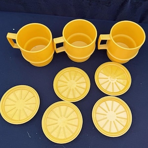 May include: Set of three yellow ceramic mugs with matching yellow coasters. The mugs have a rounded shape and a smooth finish. The coasters have a circular shape with a sunburst design.