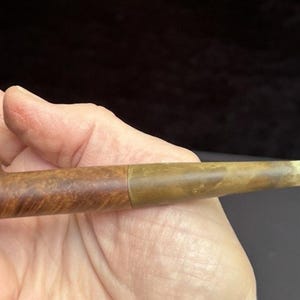 Pode incluir: Um cachimbo de tabaco vintage com uma piteira e um fornilho de madeira. O fornilho é castanho escuro, e a piteira é castanha clara com uma faixa de latão. O cachimbo é segurado em uma mão.