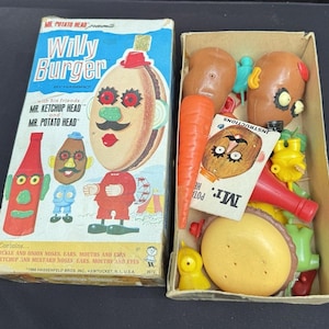 Könnte beinhalten: Vintage Mr. Potato Head "Willy Burger" Spielzeugset in der Originalverpackung. Die Schachtel zeigt Illustrationen der Charaktere und den Text "Mr. Potato Head". Die offene Schachtel zeigt verschiedene Kunststoff-Spielzeugteile, darunter eine Karotte, einen Burger und Kartoffelkopf-Teile.