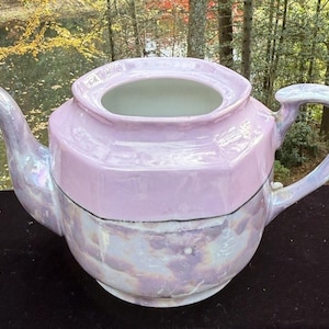 May include: A pearlescent pink teapot with a geometric design. The teapot has a curved spout and a large handle. The top of the teapot is open, revealing the interior. The teapot is sitting on a black surface.