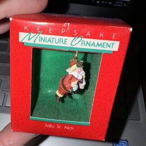 May include: A red and white Hallmark Keepsake Miniature Ornament featuring a jolly Santa Claus holding a bag of gifts. The ornament is in a clear plastic display box with a red background and the text "Keepsake Miniature Ornament" and "Jolly St. Nick".