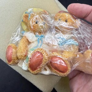 May include: Two small, tan-colored teddy bear figurines, each wearing a bonnet and dress with blue accents. The bears are in a seated position, with visible pink paws. The figurines are packaged in clear plastic.