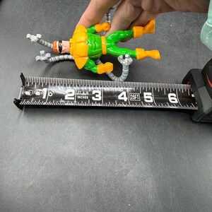Puede incluir: Una figura de acci&oacute;n del Dr. Octopus, con un traje verde y naranja, se mide con una cinta m&eacute;trica. La figura mide aproximadamente 10 cm de largo. La figura tiene cuatro brazos mec&aacute;nicos plateados.