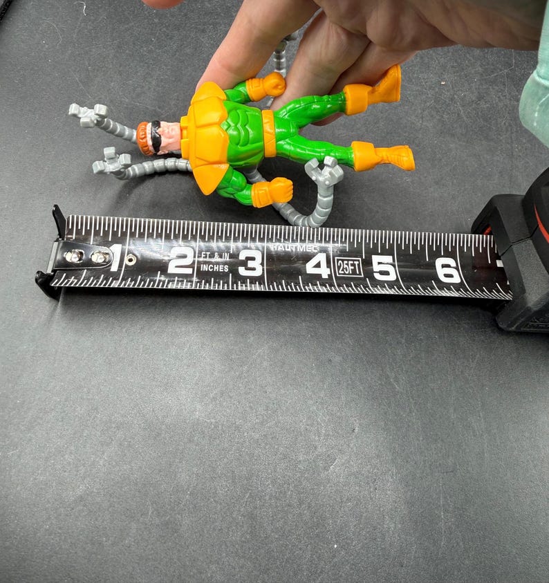 Puede incluir: Una figura de acci&oacute;n del Dr. Octopus, con un traje verde y naranja, se mide con una cinta m&eacute;trica. La figura mide aproximadamente 10 cm de largo. La figura tiene cuatro brazos mec&aacute;nicos plateados.